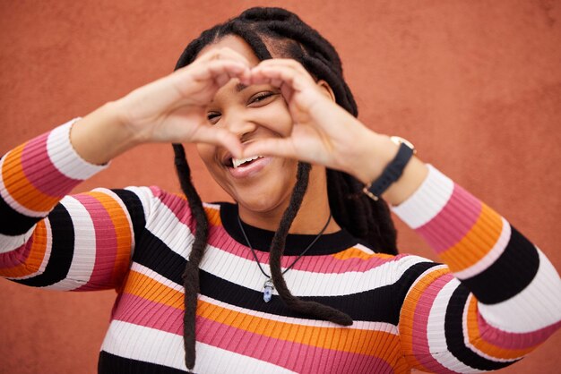 Foto forma de corazón feliz y mujer negra junto a una pared en la ciudad mientras caminaba en un viaje de fin de semana retrato de felicidad y mujer africana con un emoji de amor o gesto de mano en la ciudad de vacaciones o vacaciones