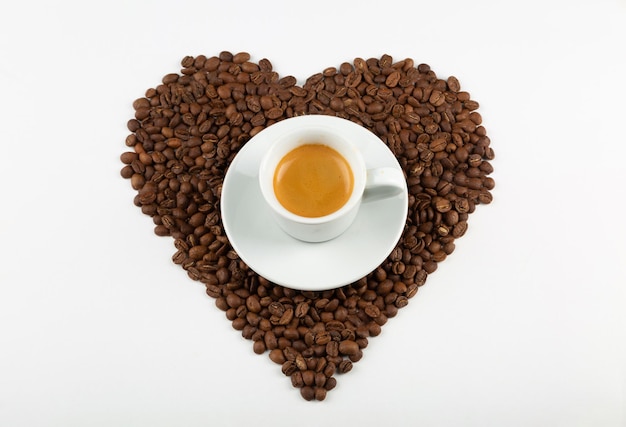 Forma de corazón hecha de granos de café y una taza de café capuchino sobre un fondo blanco.