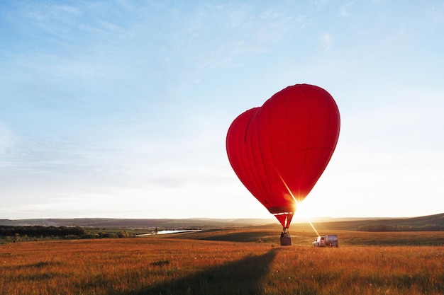 Forma de coração de balão de ar vermelho quente voando ao pôr do sol sobre o vale, pousando ou decolando.