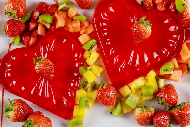 Forma de coração de gelatina vermelha com frutas frescas picadas em uma mesa branca