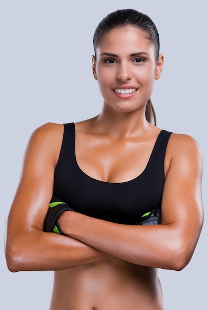 En forma y hermosa. Hermosa joven deportiva en ropa deportiva manteniendo los brazos cruzados y sonriendo mientras está de pie contra el fondo gris