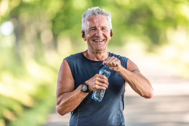 En forma sonriente y hombre jubilado con cabello gris sonríe mientras abre una botella de agua después del entrenamiento