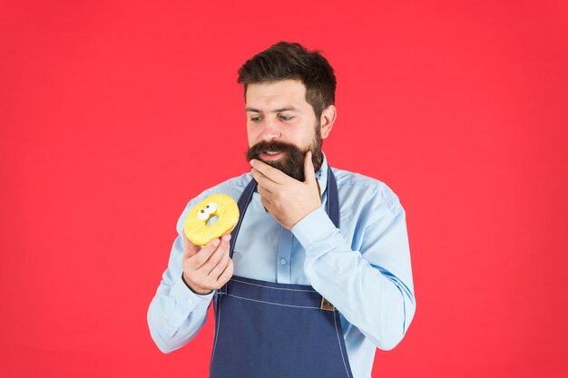 Formas de reducir el hambre y el apetito. Panadero barbudo hipster mantenga donut glaseado sobre fondo rojo. Concepto de cafetería y panadería. Donut dulce de panadero. El panadero barbudo del hombre en el delantal de cocina sostiene el postre lindo.