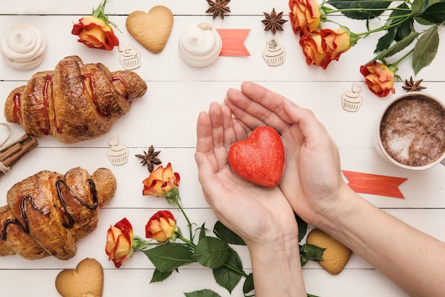 Formato de coração nas mãos sobre a mesa decorada para o Dia dos Namorados, croissants frescos e flores