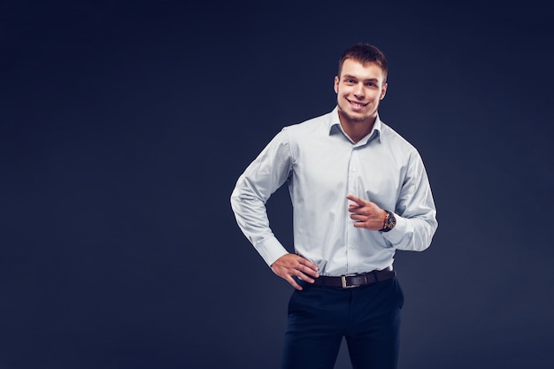 Forme al hombre serio joven en camisa pelada señala el dedo, sonriendo y mirando la cámara en fondo oscuro.