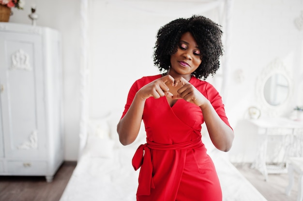 Forme a modelo afroamericano en el vestido rojo de la belleza, mujer atractiva que presenta el vestido de noche en el sitio blanco de la vendimia y muestre el corazón por las manos.