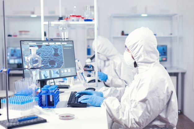 Forscher, die Informationen eingeben, die in einem modernen Labor erhalten wurden. Medizintechniker, der Computer während der globalen Pandemie mit Coronavirus im Overall verwendet.
