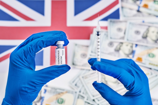 Forscherhand in den blauen Handschuhen, die Coronavirus, Covid-19-Impfstoff gegen Hintergrund der Flagge von England und Geldkrankheit halten, die für Impfschuß der klinischen Studien des Menschen, Medizinkonzept vorbereiten.