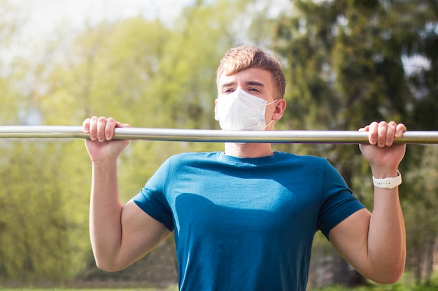 Foto forte ajuste cara, jovem atlético na máscara protetora médica fazendo esporte exersice, pull-up na barra horizontal, treino ao ar livre durante a quarentena. estilo de vida saudável, coronavírus, conceito de covid-19