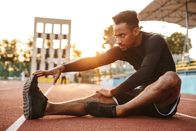 Forte homem afro-americano esticando o corpo enquanto está sentado em um campo esportivo ao ar livre