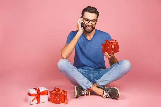 Foto adorable del hombre joven atractivo con la sonrisa hermosa que sostiene las cajas del presente de cumpleaños aisladas sobre la pared rosada, sentándose en el piso. Concepto de regalo. Usando el teléfono.