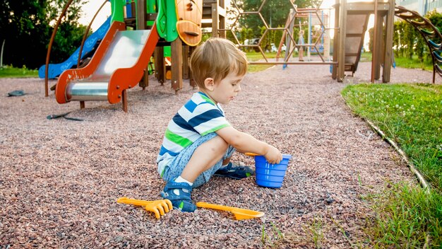 Foto de adorable niño de 3 años sentado en el patio de recreo y cavando arena con una pequeña pala de plástico y un balde
