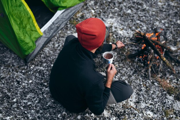 Foto aérea ao ar livre do jovem macho bebendo bebida quente nas montanhas perto da fogueira Vista traseira do viajante de chapéu vermelho sentado perto da barraca de acampamento segurando uma caneca de chá após caminhadas Viagens