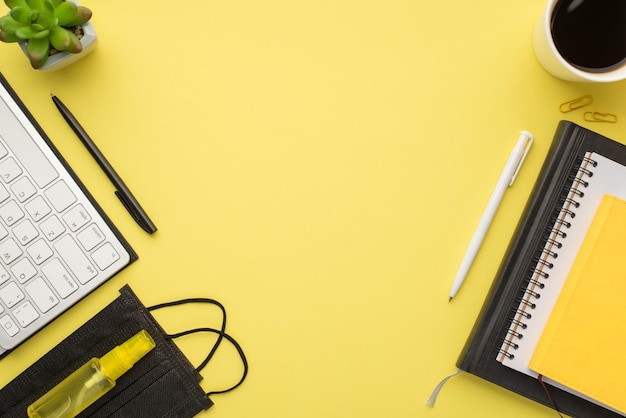 Foto aérea da planta da caneta do teclado xícara de café clipes de papel máscara de notebook e desinfetante isolado no fundo amarelo