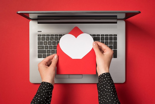 Foto aérea de laptop cinza e mãos segurando um envelope vermelho com coração de papel branco dentro isolado no pano de fundo vermelho