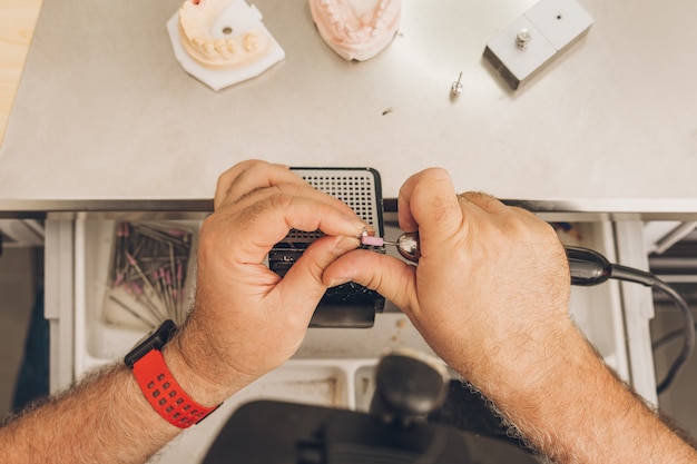 Foto aérea de las manos de un varón adulto de raza caucásica limar con un molde de cerámica con una herramienta eléctrica en un laboratorio dental especializado en cerámica ósea dental para crear coronas de porcelana