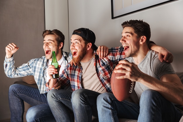 Foto de aficionados al fútbol gritando alegremente gol, mientras ven el partido y beben cerveza en compañía masculina en casa