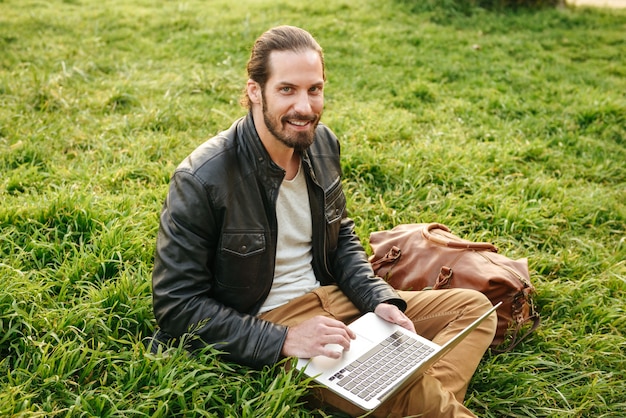 Foto al aire libre de un chico guapo con estilo en chaqueta de cuero mirándote, mientras está sentado en el césped en el parque verde y usa un cuaderno plateado