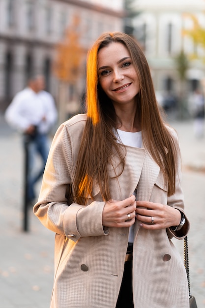 Foto al aire libre de una dama morena caminando sobre el fondo de la calle en día de otoño. Retrato de estilo callejero de moda. vistiendo pantalones casuales oscuros, suéter blanco y abrigo color crema. Concepto de moda.