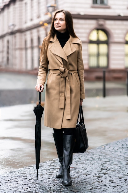 Foto al aire libre de una dama morena posando con paraguas negro en un día lluvioso de otoño. Retrato de estilo callejero de moda. vistiendo pantalones casuales oscuros, suéter blanco y abrigo color crema. Concepto de moda.