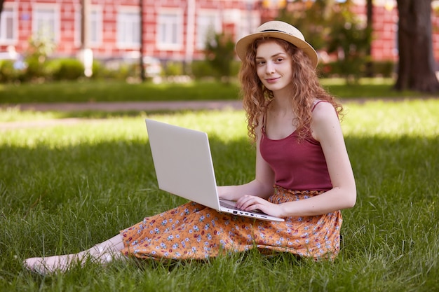 Foto al aire libre de la guapa modelo encantadora posando sobre el césped en el parque, vistiendo falda colorida, top rojo y sombrero de paja, mirando a un lado, con una sonrisa sincera en la cara, sosteniendo su dispositivo con ambas manos.