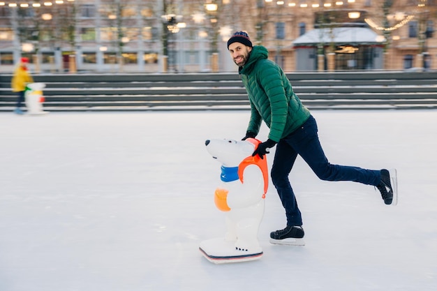 La foto al aire libre de un hombre feliz y encantador con barba espesa y bigote mira alegremente a la cámara y demuestra ir a patinar en un anillo de hielo con ayuda para patinar contento de practicar deporte de esa manera