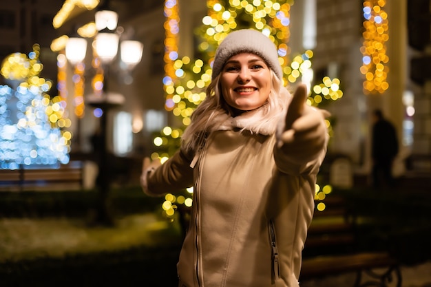 Foto al aire libre de la joven hermosa niña sonriente feliz con luces de bengala, posando en la calle. Feria de Navidad festiva en el fondo. Modelo vistiendo elegante abrigo, gorro de punto, bufanda.