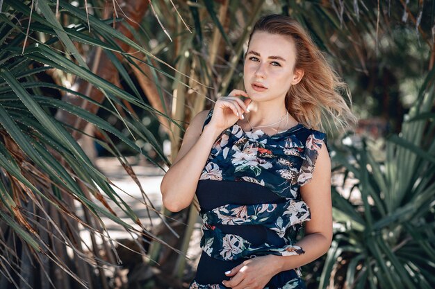 Foto al aire libre de moda de hermosa mujer sensual con cabello rubio luce lujoso vestido colorido con estampado de flores, posando en el fondo de palmeras