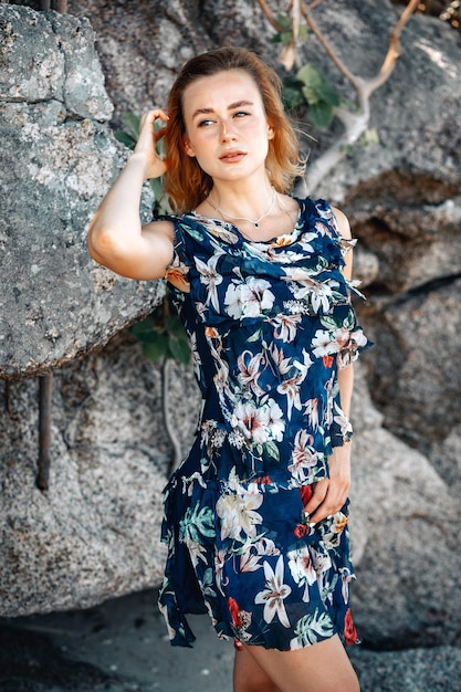 Foto al aire libre de moda de hermosa mujer sensual con cabello rubio viste lujoso vestido colorido con estampado de flores, posando en el fondo de piedras