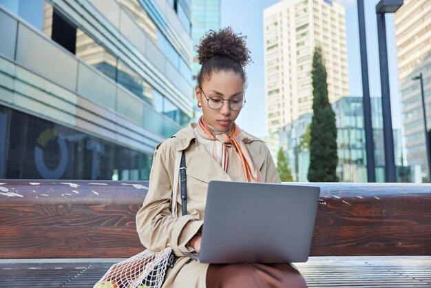 Una foto al aire libre de una mujer de cabello rizado con un abrigo elegante usa teclados de computadoras portátiles y trabaja en poses de proyectos financieros en un banco de madera contra el fondo de la ciudad moderna La directora ejecutiva trabaja a distancia