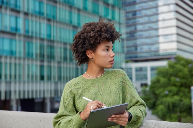 La foto al aire libre de una mujer pensativa con expresión seria usa una tableta digital y un lápiz óptico para crear dibujos, usa un puente verde casual enfocado en poses contra edificios urbanos en la ciudad