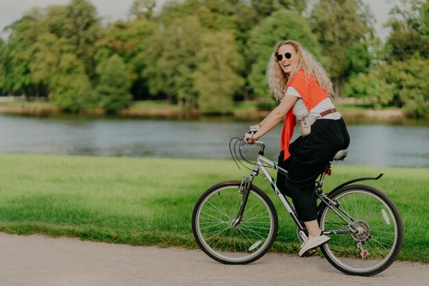 Una foto al aire libre de una mujer rubia feliz y rizada usa gafas de sol vestidas con ropa informal activa, paseos en bicicleta cerca del lago y árboles verdes en el campo, pasa tiempo libre afuera y disfruta de su pasatiempo favorito
