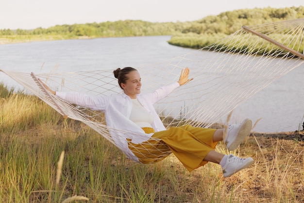 Una foto al aire libre de una mujer sonriente y satisfecha con camisa blanca sentada en una hamaca en la orilla del río extiende las manos mirando hacia otro lado disfrutando de la hermosa naturaleza