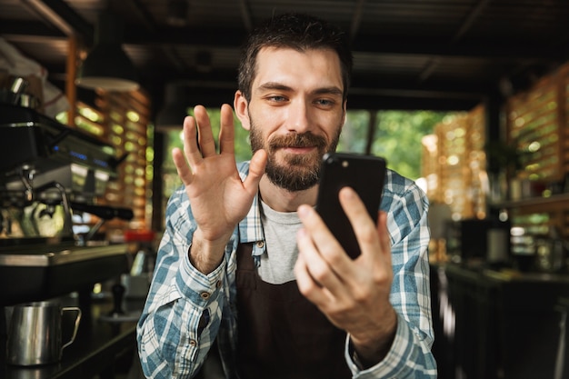 Foto de alegre chico barista vistiendo delantal sonriendo mientras usa el teléfono inteligente en la cafetería o cafetería al aire libre