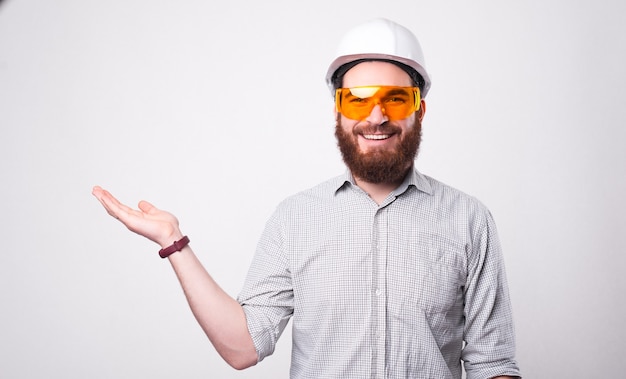 Foto de alegre guapo arquitecto hombre con casco y gafas protectoras sosteniendo copyspace en la palma