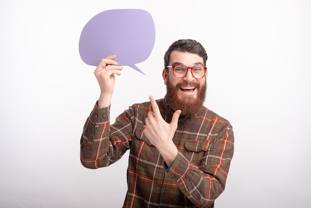 Foto de alegre joven con barba con gafas y apuntando al discurso de burbuja