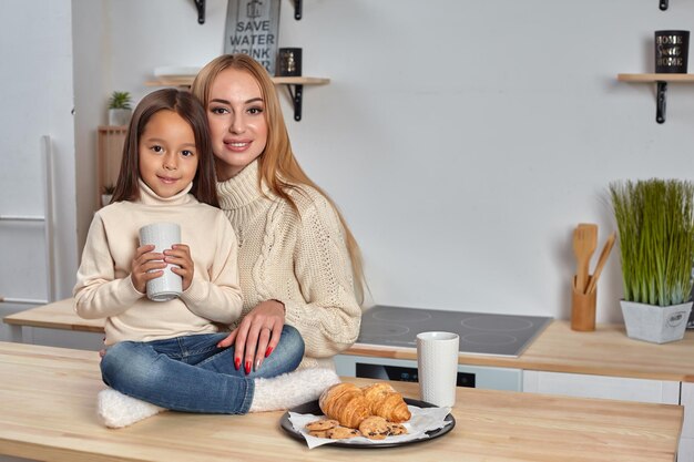 Foto de alegre madre e hija se sientan juntas en la mesa de la cocina beben té caliente en la mañana tienen ple ...