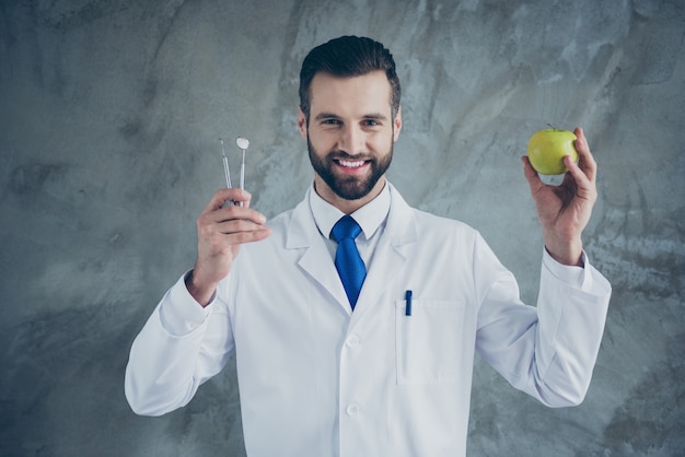 Foto de alegre médico positivo sosteniendo instrumentos y apple vistiendo bata blanca sonriendo toothily pared de hormigón de color gris aislado
