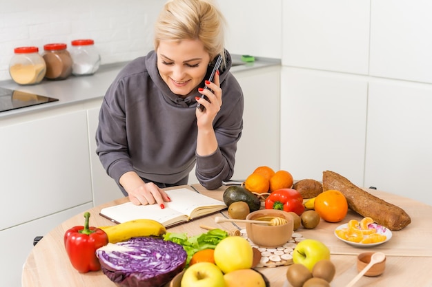 Foto de una alegre mujer caucásica sosteniendo un smartphone mientras cocina ensalada con verduras frescas en el interior de la cocina en casa.