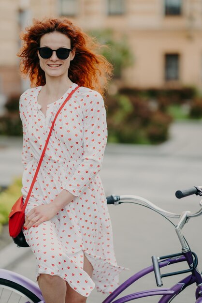 Foto de una alegre mujer pelirroja con una sonrisa amable que pasa el tiempo libre montando en bicicleta en las calles de la ciudad durante un día soleado y ventoso viste elegantes poses de vestido de verano sobre el fondo borroso de la ciudad Toma al aire libre