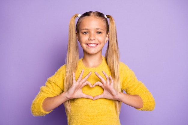 Foto de alegre niña positiva sonriendo toothily elegante moda mostrando signo de forma de corazón con los dedos aislados de pared de color violeta pastel