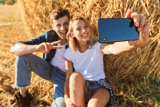 Foto de la alegre pareja hombre y mujer tomando selfie en smartphone mientras está sentado bajo un gran pajar en campo dorado, durante el día soleado