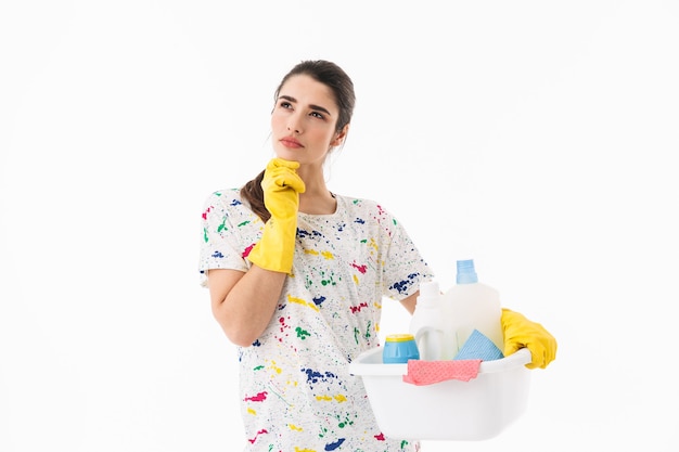 Foto de ama de casa adorable de 20 años con guantes de goma amarillos para la protección de las manos sosteniendo un balde con suministros de limpieza aislado sobre una pared blanca