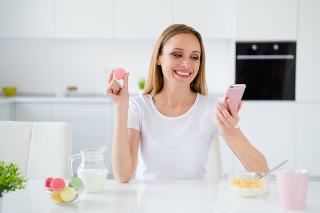 Foto de ama de casa bonita sosteniendo teléfono charlando con amigos comiendo macarrones coloridos granola desayuno leche en la mesa de luz blanca cocina interior