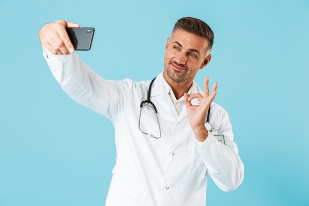 Foto de amable médico con bata blanca y un estetoscopio tomando selfie en smartphone, que se encuentran aisladas sobre la pared azul