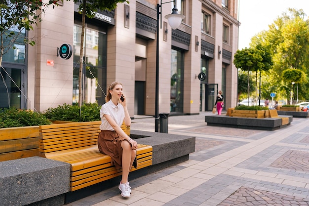 Foto ampla de uma jovem elegante com cabelos loiros conversando no celular sentada no banco