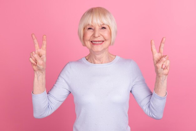 Foto de una anciana de buen humor que muestra los dedos de la paz y el símbolo fresco de viaje aislado sobre un fondo de color rosa