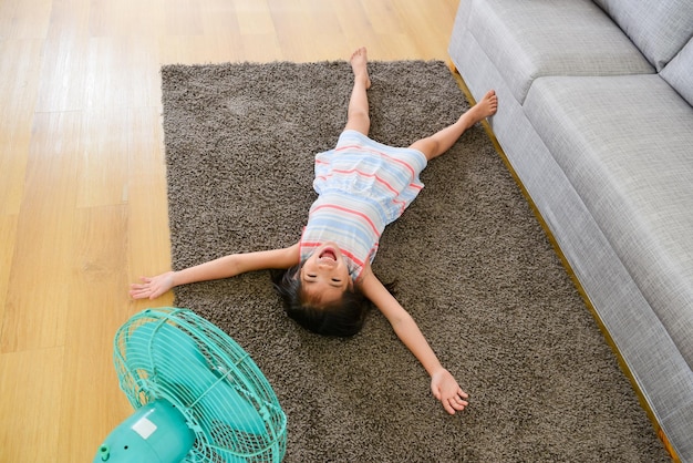 foto en ángulo alto de una niña feliz y linda acostada en el suelo y disfrutando del ventilador eléctrico que sopla viento fresco en el día de verano.