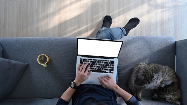 Foto anterior de un joven trabajando en una computadora portátil mientras está sentado en un cómodo sofá con su gato.