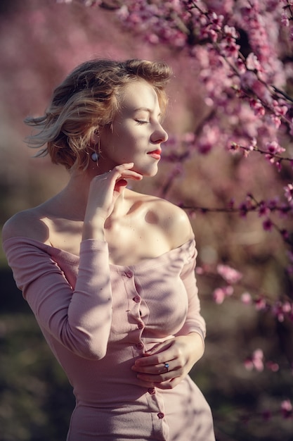 Foto ao ar livre de moda de uma jovem linda em um vestido elegante, posando no jardim com pessegueiros de flor. Loira em jardins floridos